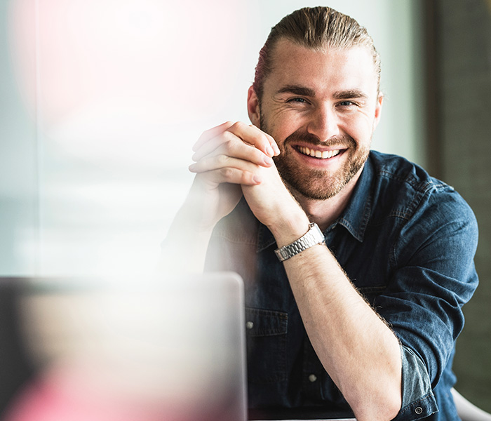 En mand ser smilende ind i kameraet. Hans computer står foran ham.